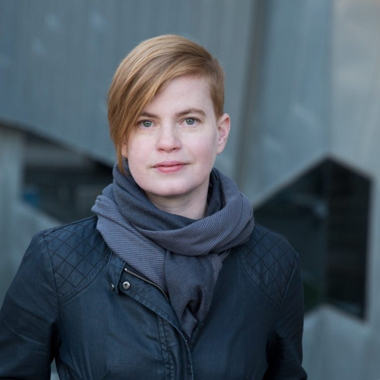 Australian composer Katharine Parton with very short red hair wearing a zip-up black jacket and dark grey scarf