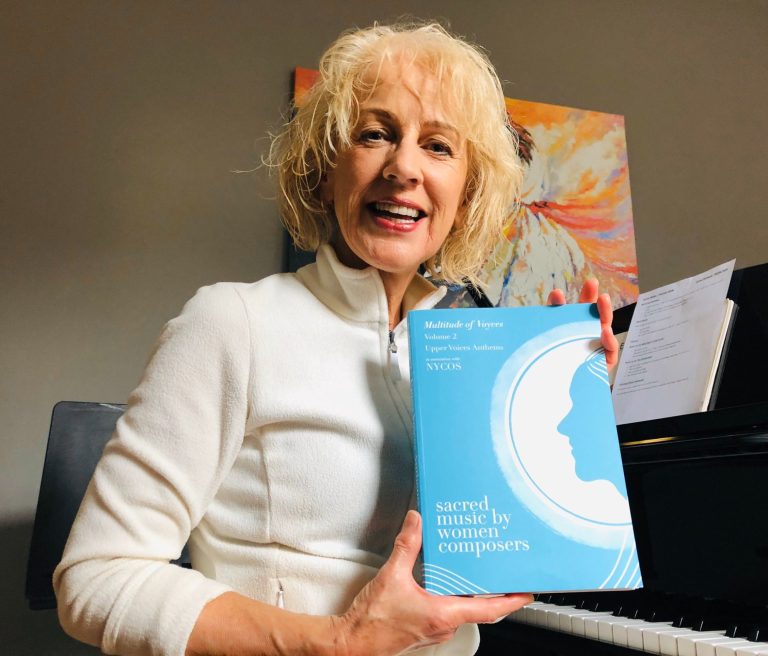 Margie Harrison - photographer unknown composer Margie Harrison with short very blonde hair wearing a white fleece sitting at her piano holding a copy of MoV Volume 2