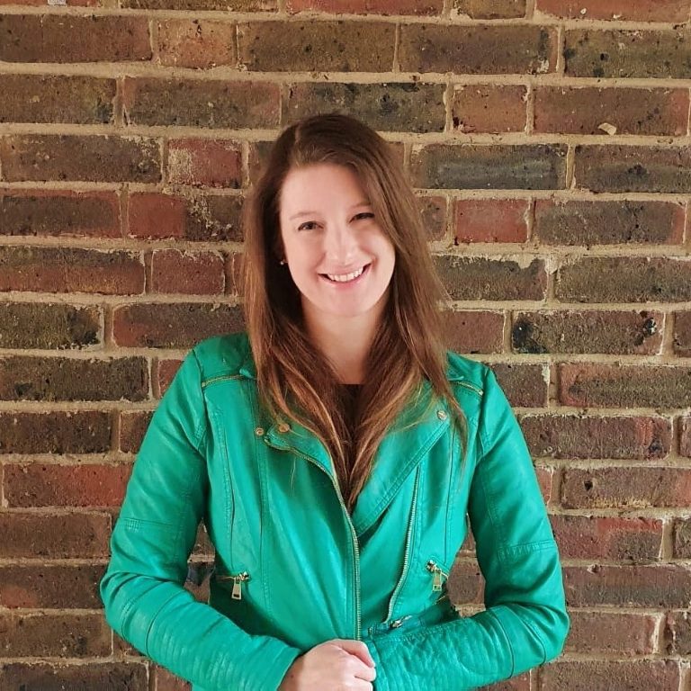 Carol J Jones - photographer unknown composer Carol J Jones with long light brown hair wearing a green leather-look jacket standing in front of a brick wall