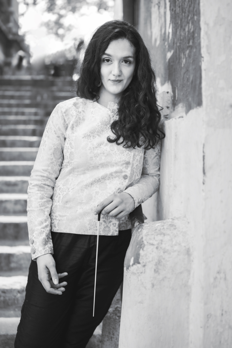 Anita Datta ©Vivienne Sarky (2021) British Asian composer Anita Datta wearing black trousers and an embroidered cream coloured jacket holding a conducting baton