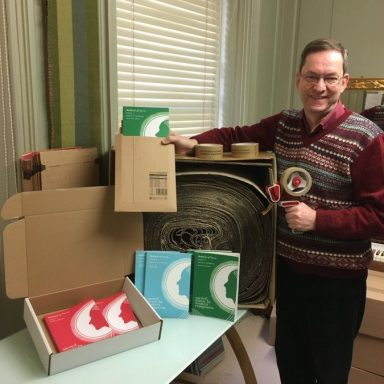 Andrew Stewart at work in the MoV office Andrew in a striped sweater stands next to boxes and anthology volumes in the office.