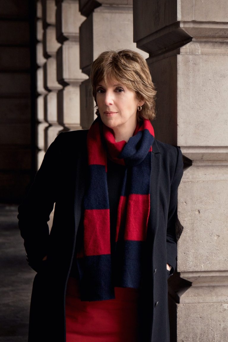 Cecilia McDowell ©Karina Lyburn composer Cecilia McDowell stands next to imposing stone pillars wearing a red skirt, blue coat and red and blue wide scarf