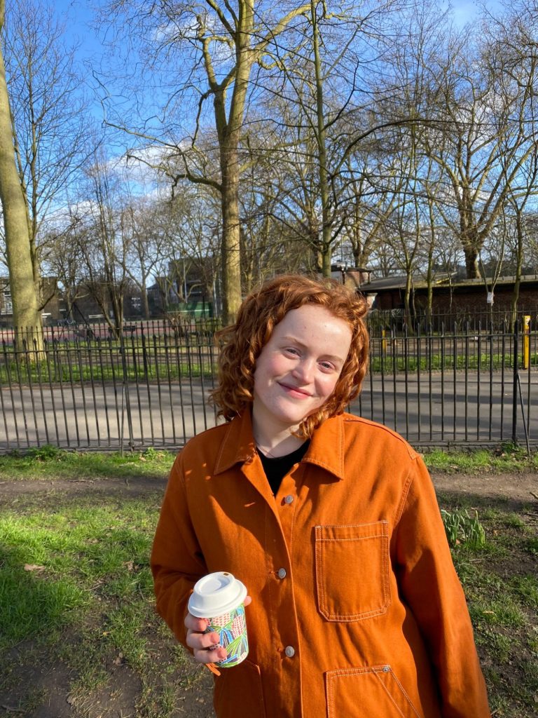young composer Joanna Ward stands in a public park with railings and trees behind. She has ginger hair and wears shirt of the same colour 