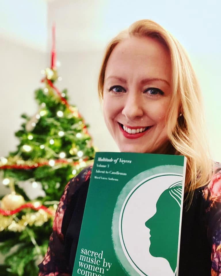 Libby Croad ©Libby Croad composer Libby Croad, with long blonde hair and multi-coloured top, holding a copy of MoV Volume 3 in front of a decorated Christmas tree