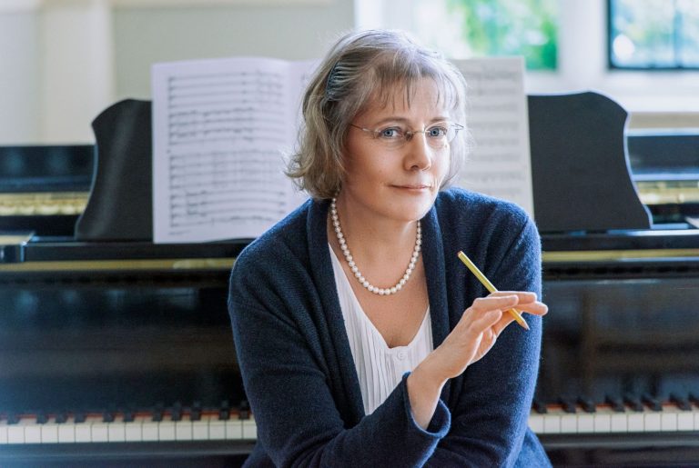 composer Janet Wheeler sits in front of a grand piano. She has a white shirt & blue cardigan. She wears a pearl necklace and holds a pencil