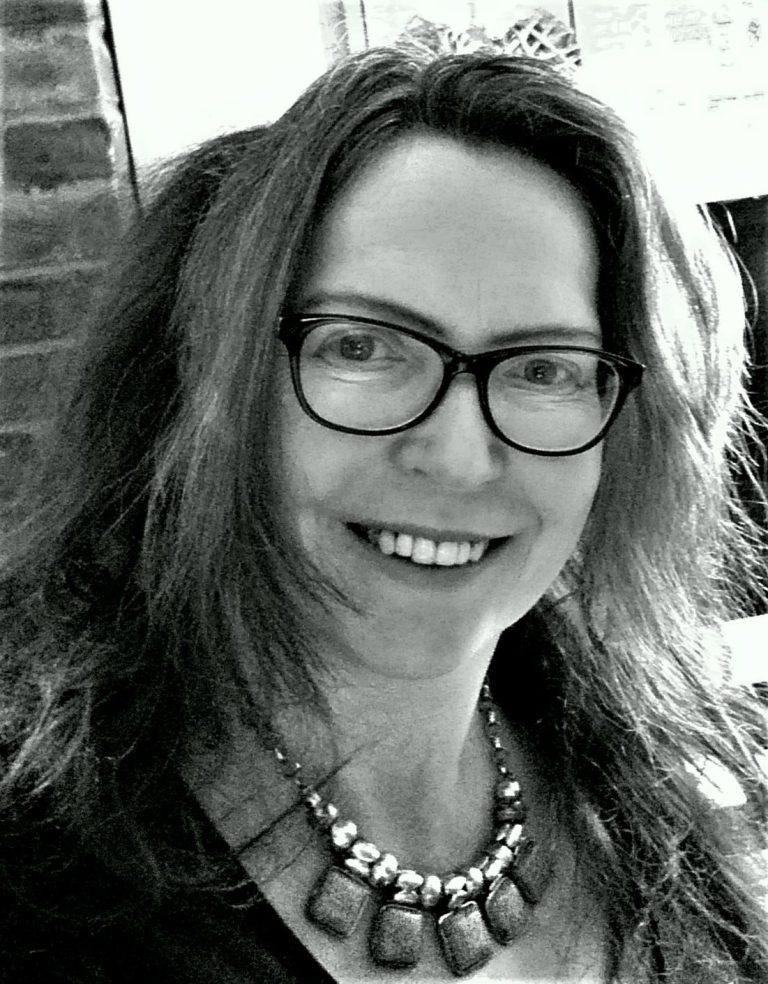 composer Alison Willis stands in the sunshine. She has a tumble of wavy brown hair and wears a modern necklace of metal and wood
