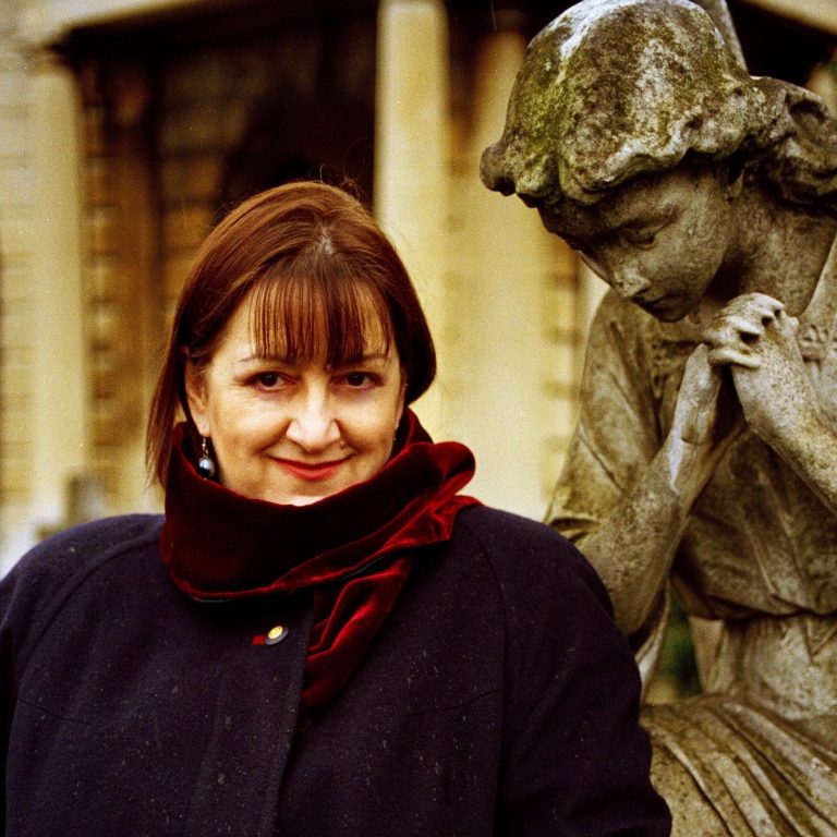 Judith Bingham OBE ©Patrick Douglas Hamilton composer Judith Bingham OBE with short chestnut brown hair wearing warm winter clothing standing by a stone statue of a female figure