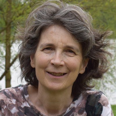 Sheena Phillips - photographer unknown composer Sheena Phillips has wavy grey brown hair, and is wearing a brown and white top. She is standing in front of tall trees in leaf