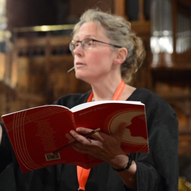 Sarah MacDonald, composer and associate Sarah reading from anthology volume 1, wearing glasses and a black robe.