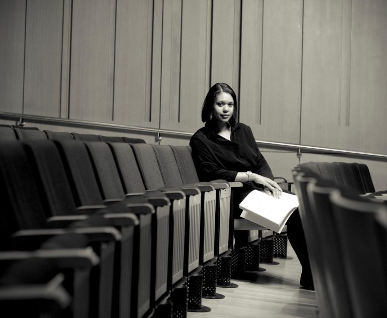 Hannah Kendall ©Kiran Ridley composer Hannah Kendall with short dark brown hair and a black dress sitting in a concert, the other seats are tipped up