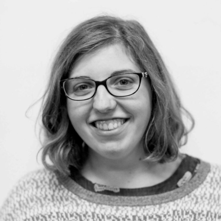 composer Sarah Cattley, with short blonde hair and glasses, wearing summery clothes, smiling broadly at the camera 