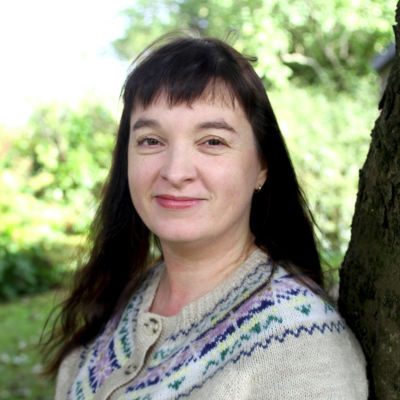 Gemma McGregor ©Scottish Music Centre composer Gemma McGregor stands in a garden with long dark brown hair wearing an intarsia wool cardigan in a traditional Scottish design