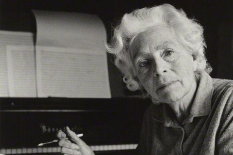 Composer Dame Elizabeth Maconchy sitting at her grand piano. On the music stand is manuscript paper; Maconchy is holding a pencil