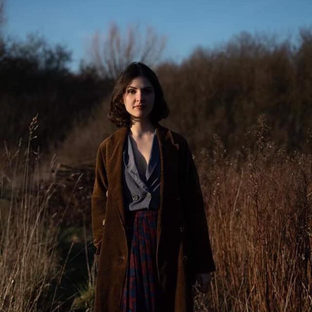 Anna Semple - photographer unknown young composer Anna Semple stands in a field of long grass wearing autumnal colours and a brown velvet coat